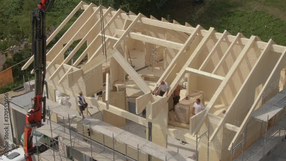 DRONE: Flying near a modern prefabricated house being built in the Slovenian countryside. Group of contractors mounts a wooden arch on the roof of a cross-laminated timber house under construction. DRONE: Flying near a modern prefabricated house being built in the Slovenian countryside. Group of contractors mounts a wooden arch on the roof of a cross-laminated timber house under construction.