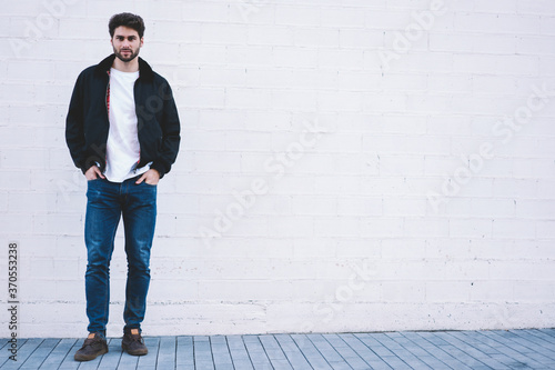 Full length portrait of stylish hipster guy standing on white promotional background for your advertising text message.Handsome man dressed in casual outfit looking at camera near copy space area
