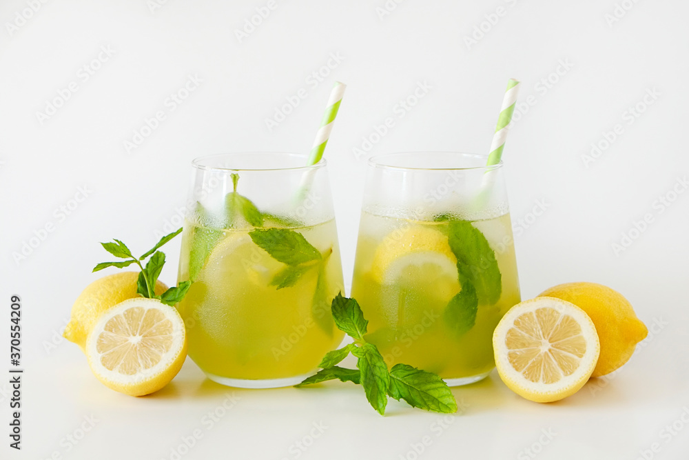 Fototapeta premium Two glasses of refreshing non alcoholic mojito drink with lemon slices, mint leaves and ice. Studio shot of iced lemonade isolated on white background, close up, copy space.