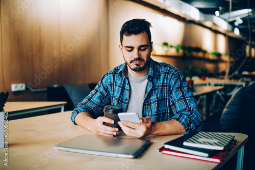 Pensive young man checking updated of friends profiles n social networks via cellular during coffee break, thinking hipster guy browsing video on telephone for watching online during leisure .