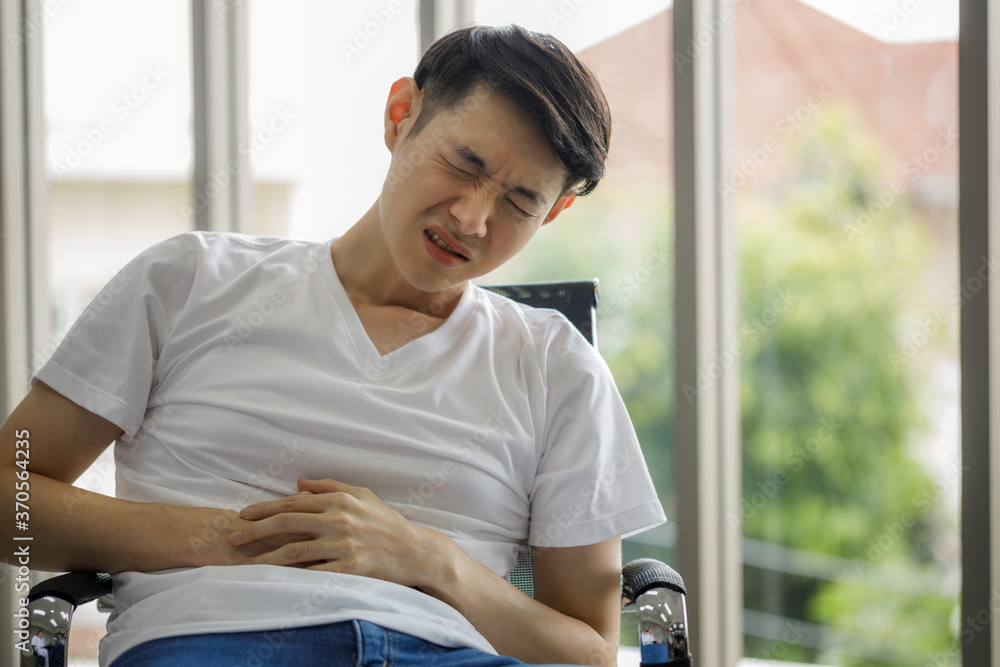 Young Asian man sits on his stomach with his hand on a chair in the home due to stomach pain from stomach ailments or stress from the effects of the COVID-19 virus.