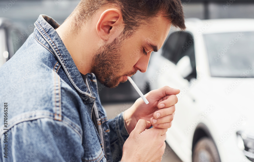 Fototapeta premium Fashionable man smoking near car outdoors against modern business building