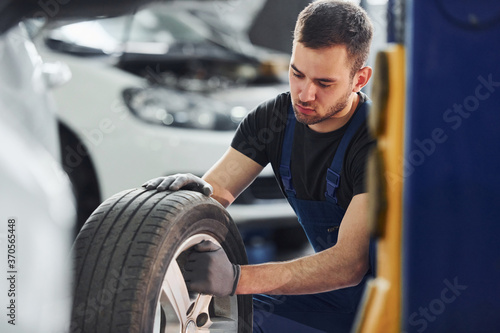 Man in work uniform sitting and changing car wheel indoors. Conception of automobile service