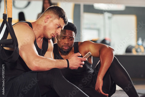 Taking a break and using phone. African american man with white guy have workout day in gym