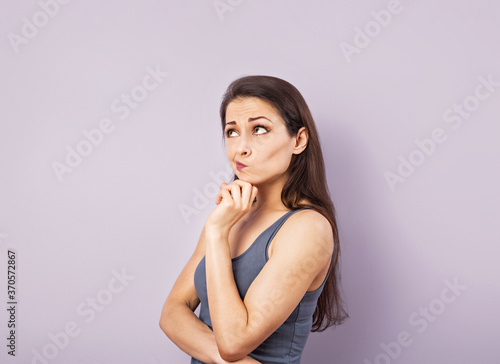 Funny grimacing brunette woman thinking and looking up in grey t-shirt with folded arms on purple background with empty copy space. Closeup