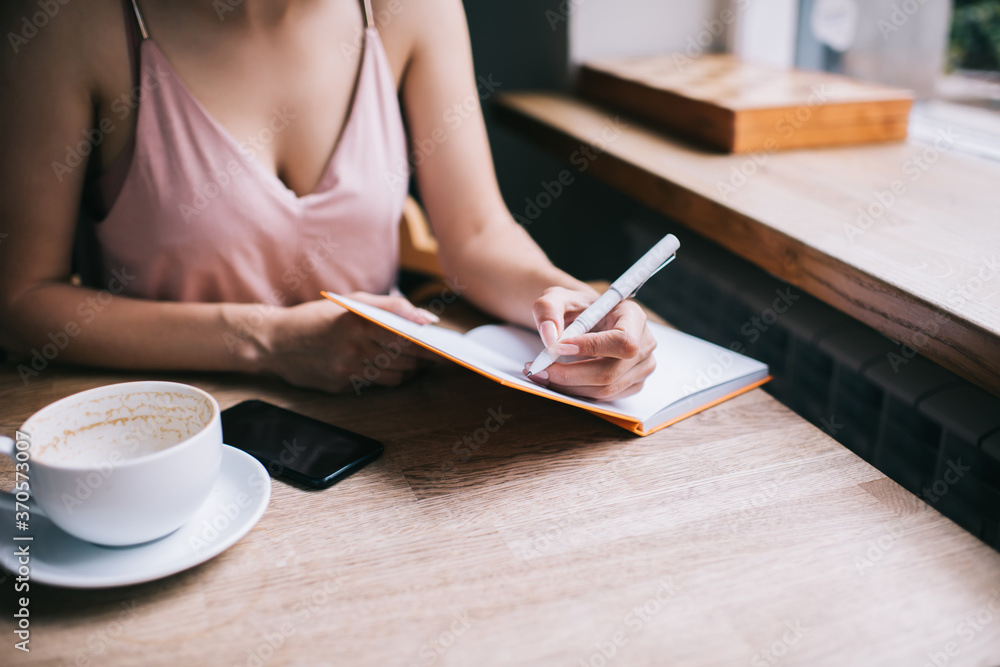 Crop feminine female writing in notebook during coffee break in modern cafeteria Crop feminine female writing in notebook during coffee break in modern cafeteria