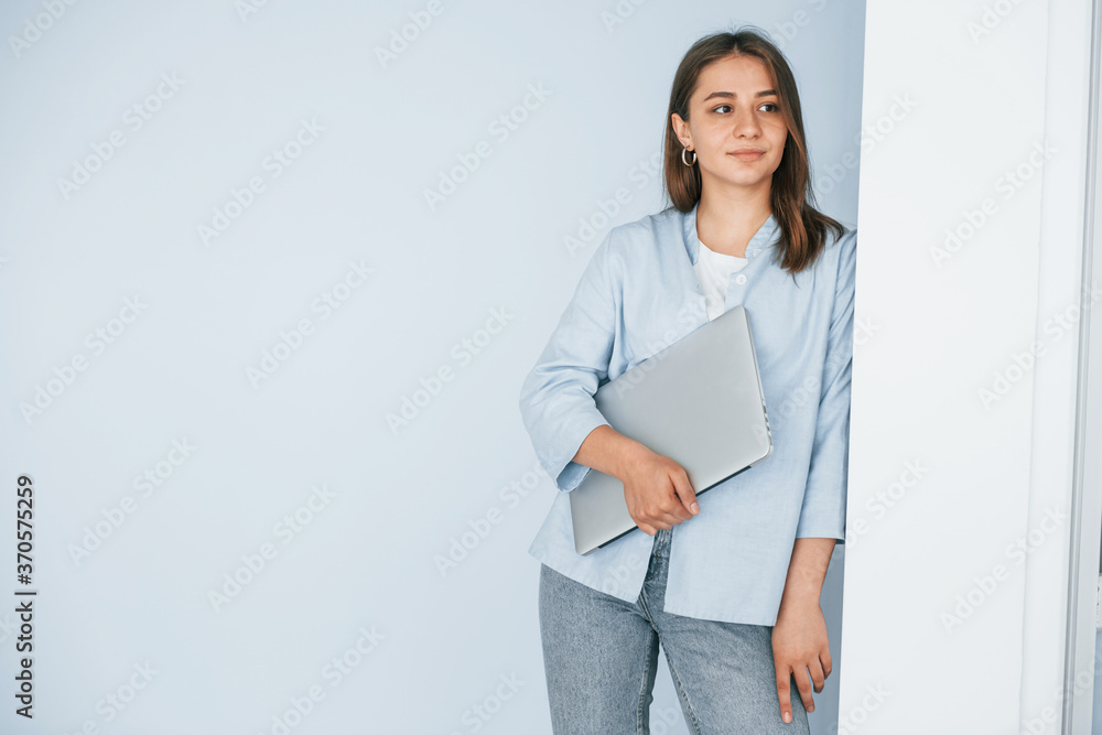 Beautiful young woman in jeans and blue shirt standing against wall with laptop in hands Beautiful young woman in jeans and blue shirt standing against wall with laptop in hands