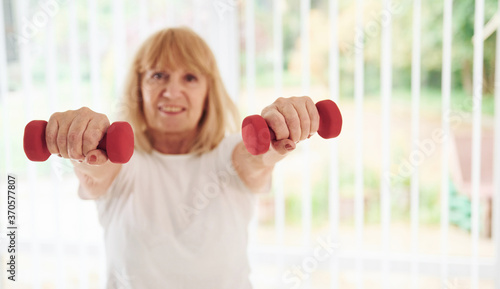 Positive senior woman in sportive clothes indoors at daytime doing exercises with dumbbells