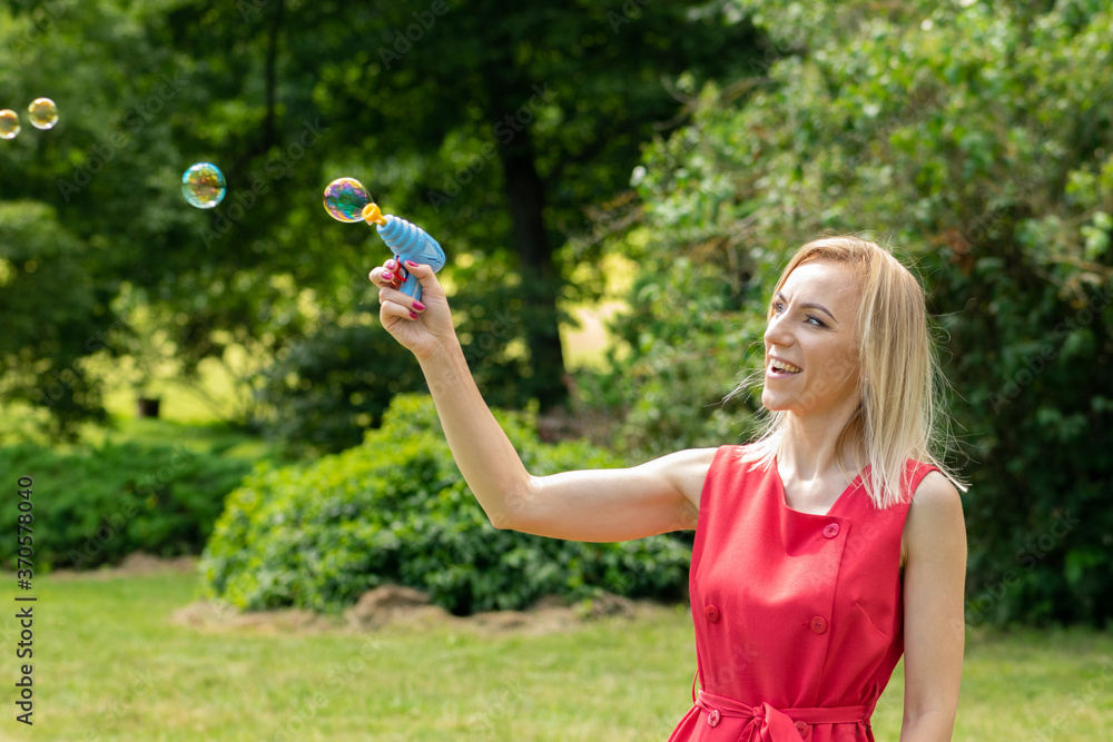 Blonde woman with red dress playing with soap bubble pistol toy during warm summer day in foliage background with copy space Blonde woman with red dress playing with soap bubble pistol toy during warm summer day in foliage background with copy space