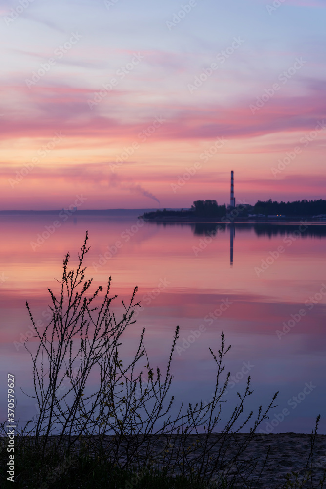 Sunset on the lake in spring with unblown bushes in the foreground and with a boiler chimney in the background