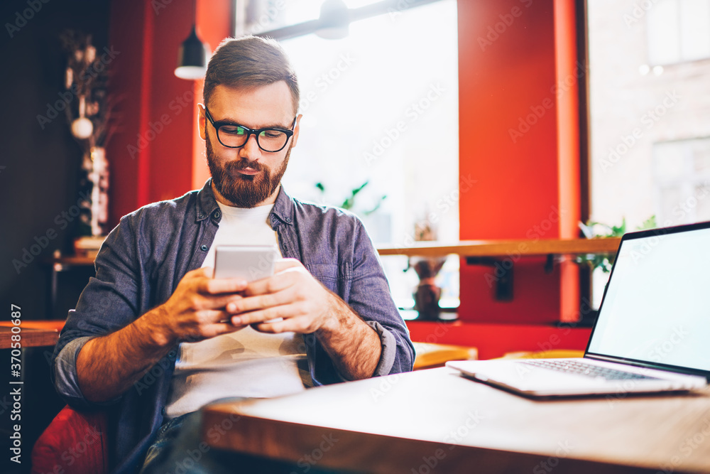 Serious male blogger in eyewear for vision correction reading sms with notification for app on smartphone device while spending time in cafe interior near table with laptop computer with mock up Serious male blogger in eyewear for vision correction reading sms with notification for app on smartphone device while spending time in cafe interior near table with laptop computer with mock up