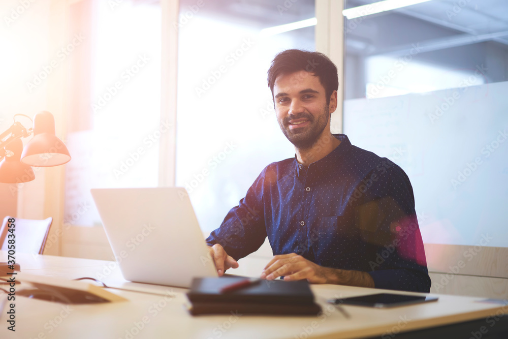 Half length portrait of handsome smiling graphic designer looking at camera while creating new model using application on laptop.Young skilled employee working with modern device sitting indoors Half length portrait of handsome smiling graphic designer looking at camera while creating new model using application on laptop.Young skilled employee working with modern device sitting indoors
