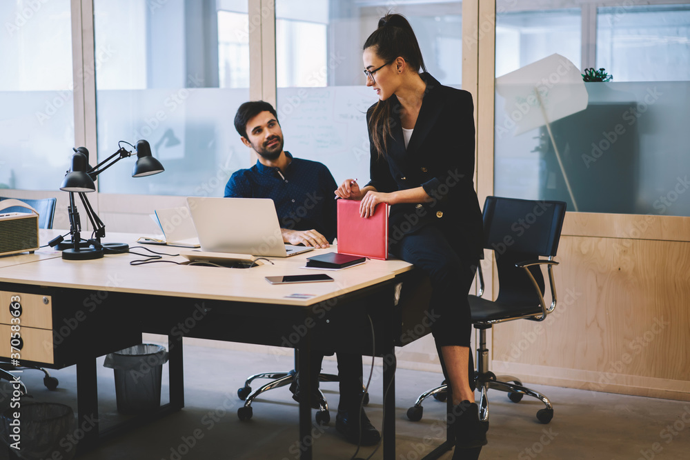 Obraz premium Attractive experienced brunette manager in spectacles sitting on wooden table and explaining main concepts of work to new talented male colleague.Coworkers communicating with each other indoors