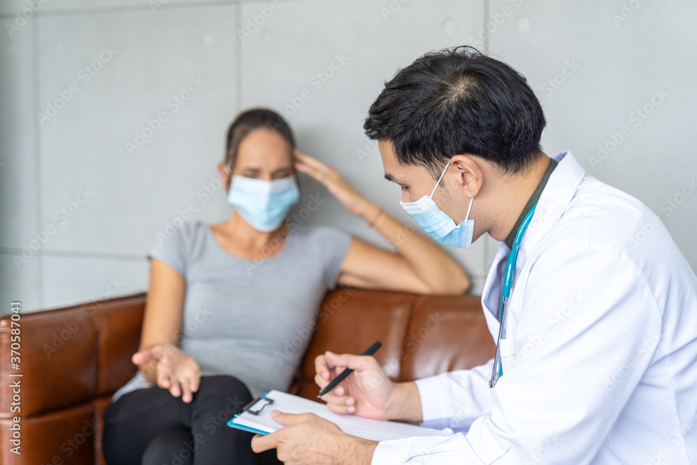 Woman wearing face mask sitting on the sofa and talking to the professional psychologist while wearing face mask conducting a consultation and making notes during coronavirus or COVID 19 outbreak