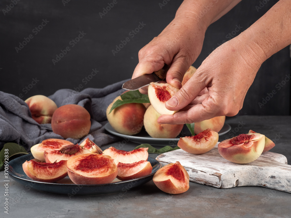The woman cuts a peach slice with a knife. There are many peaches on the table.