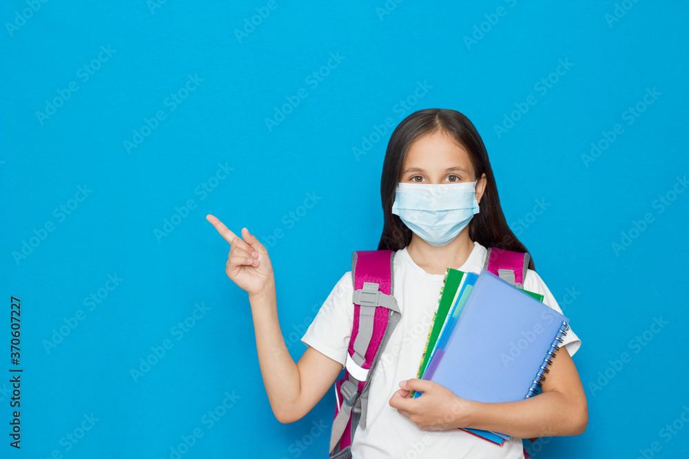 A brunette girl in a medical mask, blue background while studying at school during the pandemic advises to give feedback, choose, decide, news, information, direct, school sale.