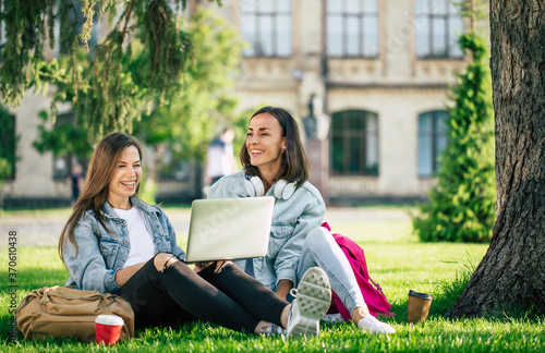 Two happy beautiful young student girl friends in casual denim clothes are relaxing in college park with laptop and smartphone on university background and drinking coffee.