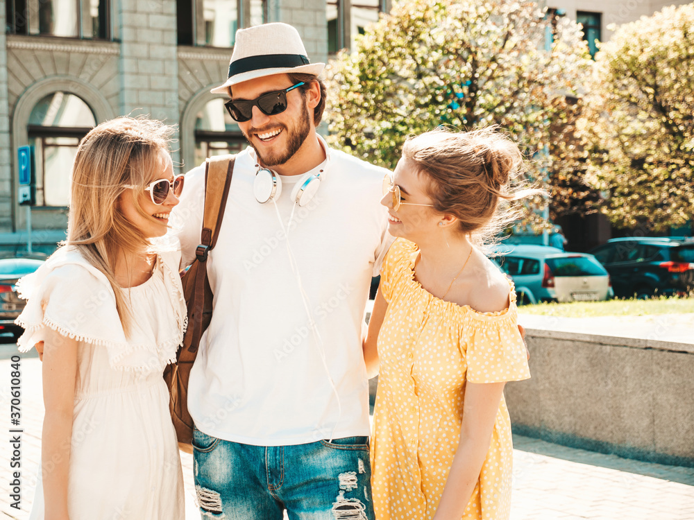 Group of young three stylish friends posing in the street. Fashion man and two cute girls dressed in casual summer clothes. Smiling models having fun in sunglasses.Cheerful women and guy chatting