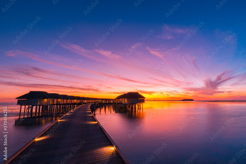 Obraz premium Sunset on Maldives island, luxury water villas resort and wooden pier. Beautiful sunset sky and clouds and tropical beach background for summer vacation holiday and travel concept banner 
