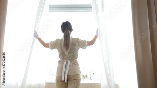 Cleaning concept in hotel or private apartament. Back view of professional hotel maid opening curtains in bright room. Pretty woman preparing accommodations for visitors at resort. 4 k video