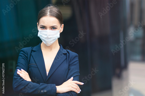 Beautiful serious business woman standing outdoors with her hands crossed, young gorgeous pretty girl in medical sterile protective mask against coronavirus, epidemic covid-19. Healthcare at job, work