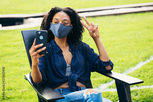 Black woman sitting in patio chair  with a face mask taking a selfie during covid 19 photo