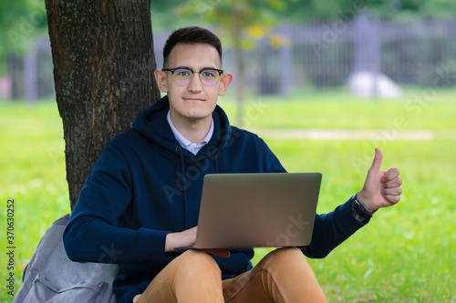 Successful happy young man, freelancer in glasses with laptop sitting near tree, leaning at wood, show thumb up, like gesture. Positive young guy working at computer outdoors. Freelance, distant job