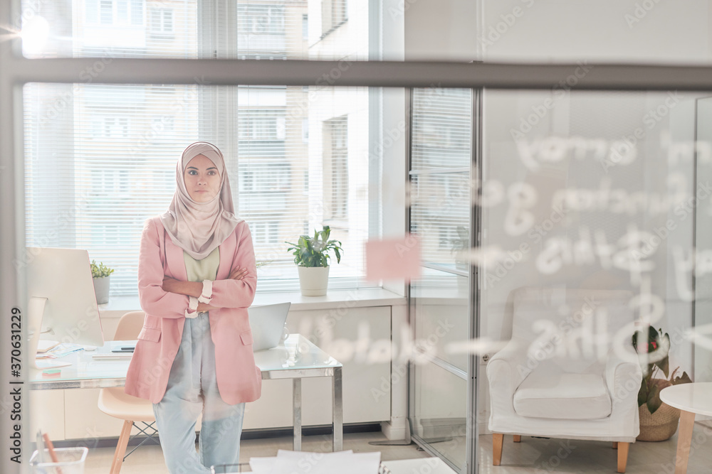 Serious young Muslim coder in hijab standing with crossed arms in office and looking at board while thinking about new app Serious young Muslim coder in hijab standing with crossed arms in office and looking at board while thinking about new app