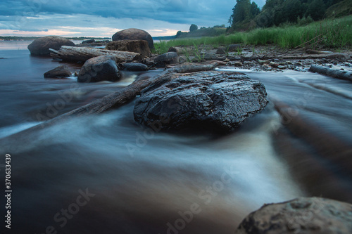 Blue mistycal night landscape with water stream in motion blur and stones in the coast. Northern Dvina river, Russia.