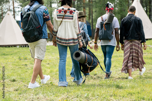 Rear view of multi-ethnic tourists in summer outfits walking on grass to tents while planning to stay in camp, festival concept