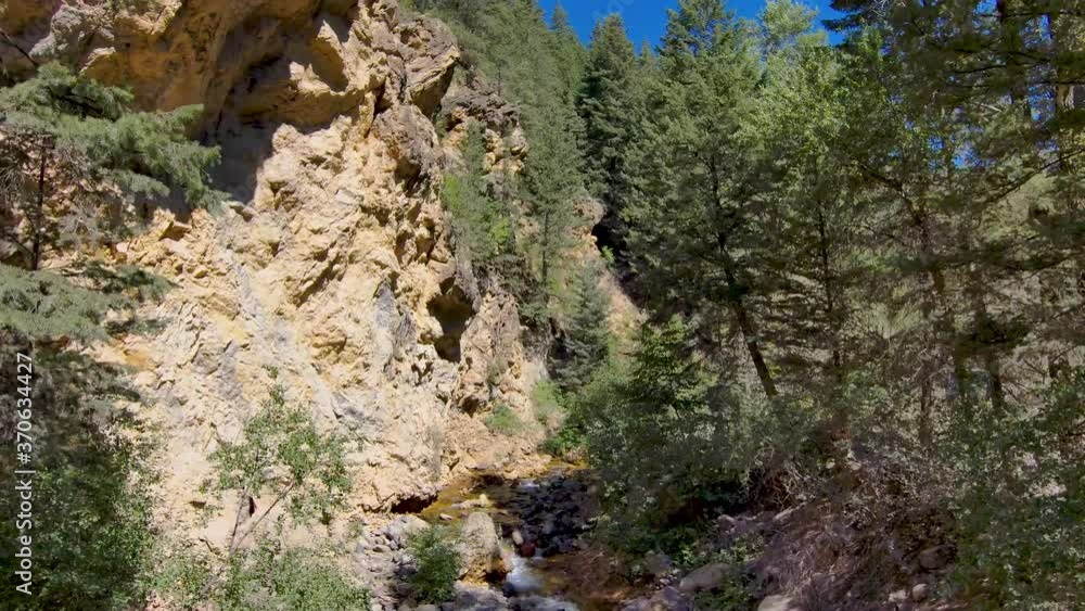 Drone flies from a stream flowing through a rugged canyon through the forest and to snow-capped peak high above the brook Drone flies from a stream flowing through a rugged canyon through the forest and to snow-capped peak high above the brook