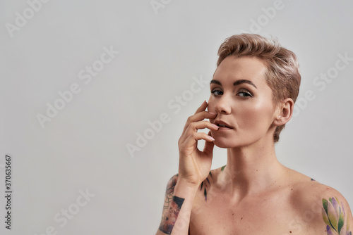 Beauty portrait of a young attractive half naked tattooed woman with perfect skin looking at camera, holding hand near her face isolated over grey background