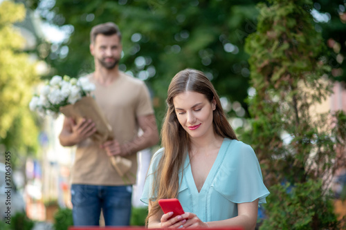 Cute female sitting, checking her phone, bearded male coming to her with flowers