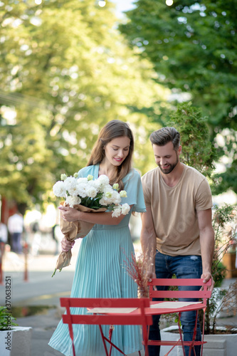 Bearded male pulling red chair for female with bouquet of white flowers, helping her to sit