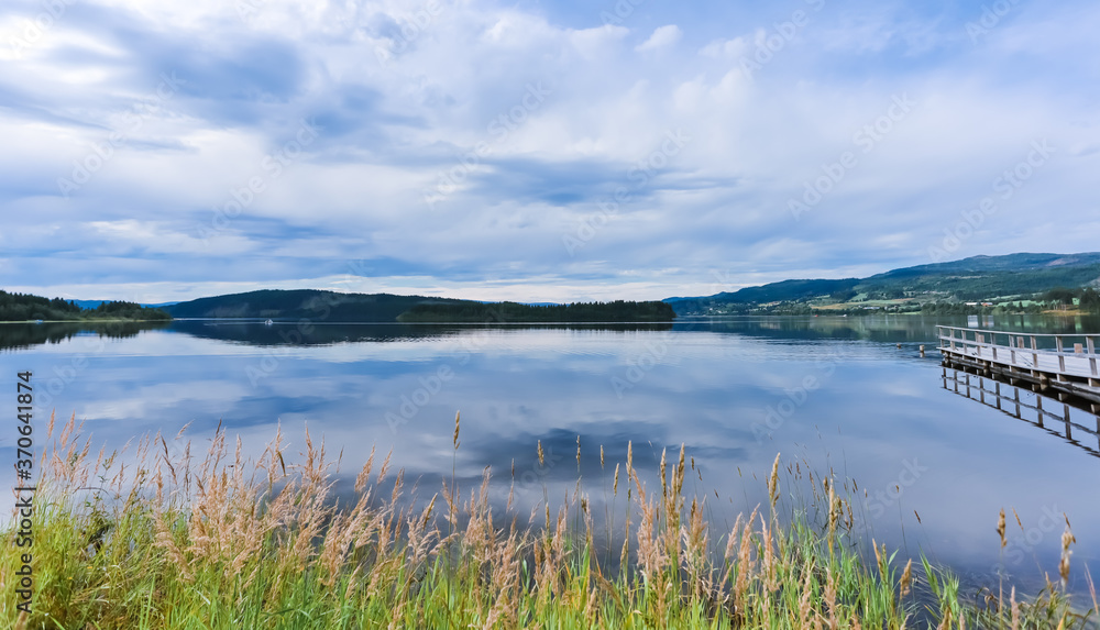 Wide angle view of the landscape, lake. The surface of Selbu Lake is tranquil. There is grass in the front, mountains in the distance, and a dock on the side.