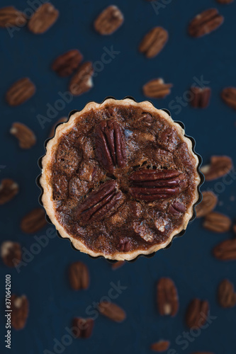 Selective focus on individual pecan pie in tart container in the foreground. Blurred out background of pecan nuts with classic dark navy blue surface. Space for copy text