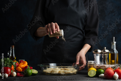 Cooking a dish of potatoes, the chef prepares potatoes for baking sprinkling with seasoning, freezing in motion, against a background of vegetables, cooking and a book of recipes