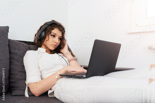 Female with curly brown hair in white outfit lying on the sofa, looking at laptop and wearing headphones.