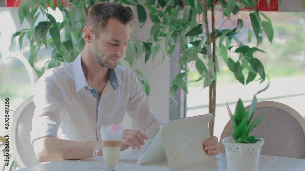 Young Customer in a Cafe Reading News Using Tablet PC. Youthful Handsome Man in a White Shirt Has a Coffee Break in the Daytime. Middle Close-Up View, Dynamic Shot Young Customer in a Cafe Reading News Using Tablet PC. Youthful Handsome Man in a White Shirt Has a Coffee Break in the Daytime. Middle Close-Up View, Dynamic Shot