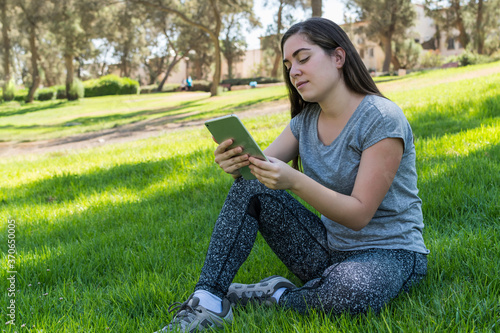 Young woman watching content on a tablet, sitting on the grass in a public park.