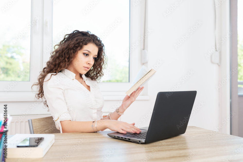Modern business woman with curly hair in white shirt sitting in office, working on laptop and holding a book. Modern business woman with curly hair in white shirt sitting in office, working on laptop and holding a book.