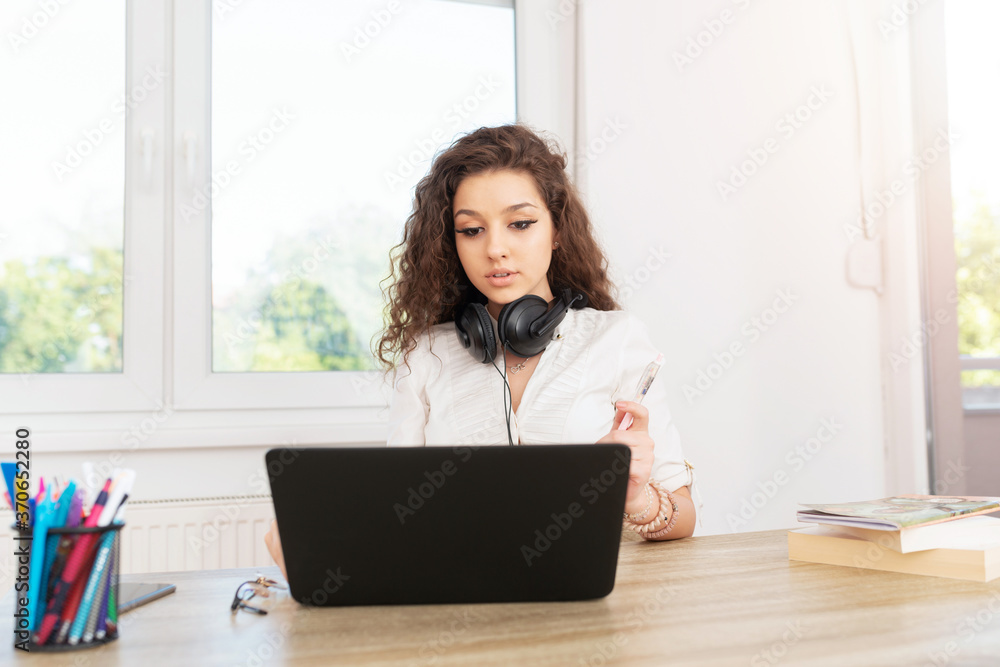 Professional young female with curly hair sitting in office by the window wearing headphones around neck and working on laptop. Professional young female with curly hair sitting in office by the window wearing headphones around neck and working on laptop.