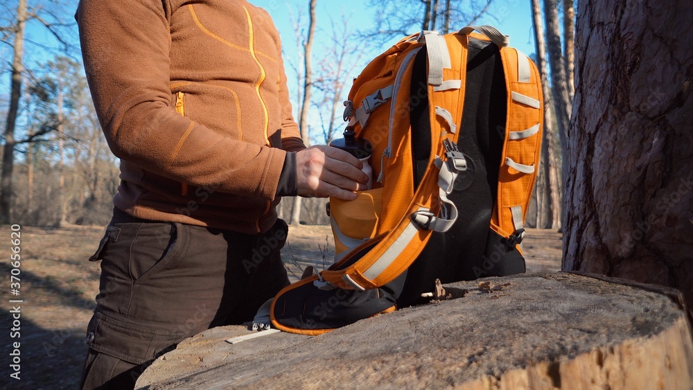Fototapeta premium Theme hiking and travel. A Caucasian tourist man unpacks an orange backpack, takes out his things and puts them on a stump in the forest. Equipment and things for camping
