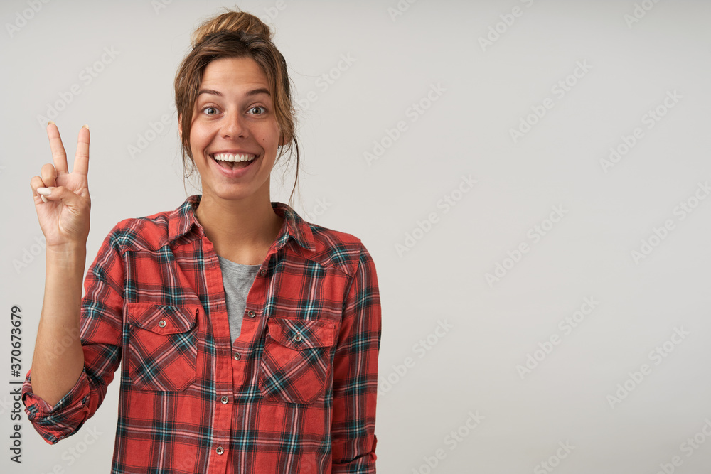 Cheerful young pretty brown haired female with natural makeup forming victory sign while looking gladly at camera with broad smile, isolated over white background Cheerful young pretty brown haired female with natural makeup forming victory sign while looking gladly at camera with broad smile, isolated over white background