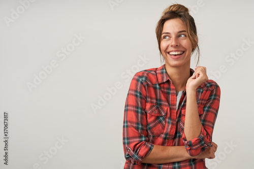 Studio shot of young pretty brown haired woman with casual hairstyle keeping her hand raised while looking happily aside with charming smile  isolated over white background