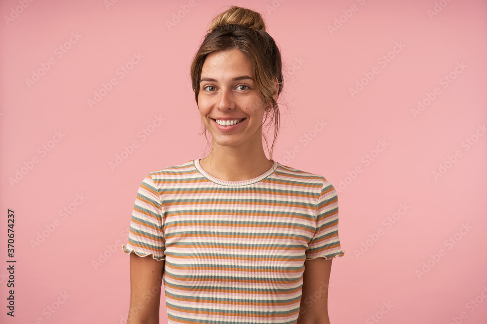 Studio photo of young lovely brown haired lady with bun hairstyle looking at camera with charming smile and keeping hands down while standing over pink background Studio photo of young lovely brown haired lady with bun hairstyle looking at camera with charming smile and keeping hands down while standing over pink background