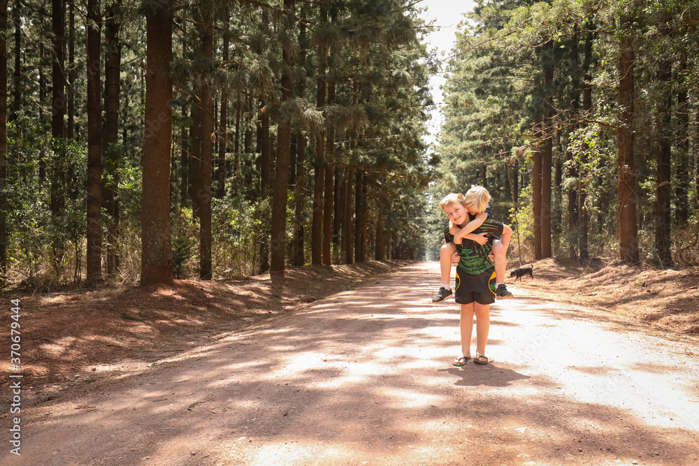 Fototapeta premium Boy giving his little brother a piggy back on dirt road among rows of tall pine trees