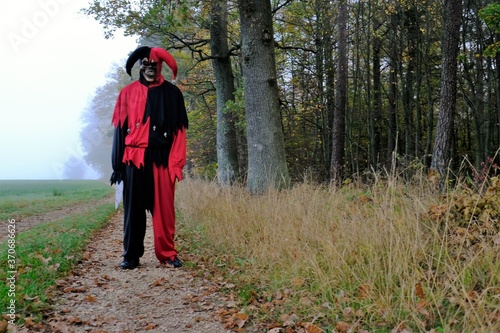 Halloween concept. Creepy clown in a skull mask on a deserted road in a foggy field near the autumn forest .Carnival in October. A man in a Evil clown costume.Autumn.Scary Jester Costume