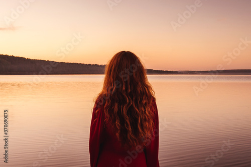 Young ginger woman in red dress at sunset. Back view. River or lake on background, pink sky.