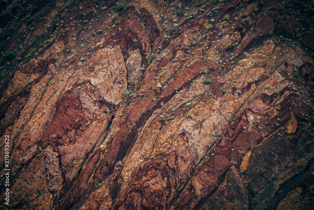 Nature background of clay mountain wall closeup. Nature texture of precipice. Natural backdrop of clay mountain surface. Magenta brown orange mountain wall with green plants close-up. Unusual slope.